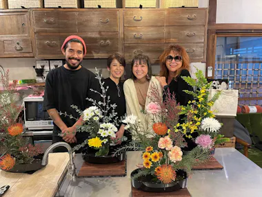 Four people stand smiling behind a table with colorful floral arrangements in a cozy room with wooden cabinets and kitchen appliances in the background.