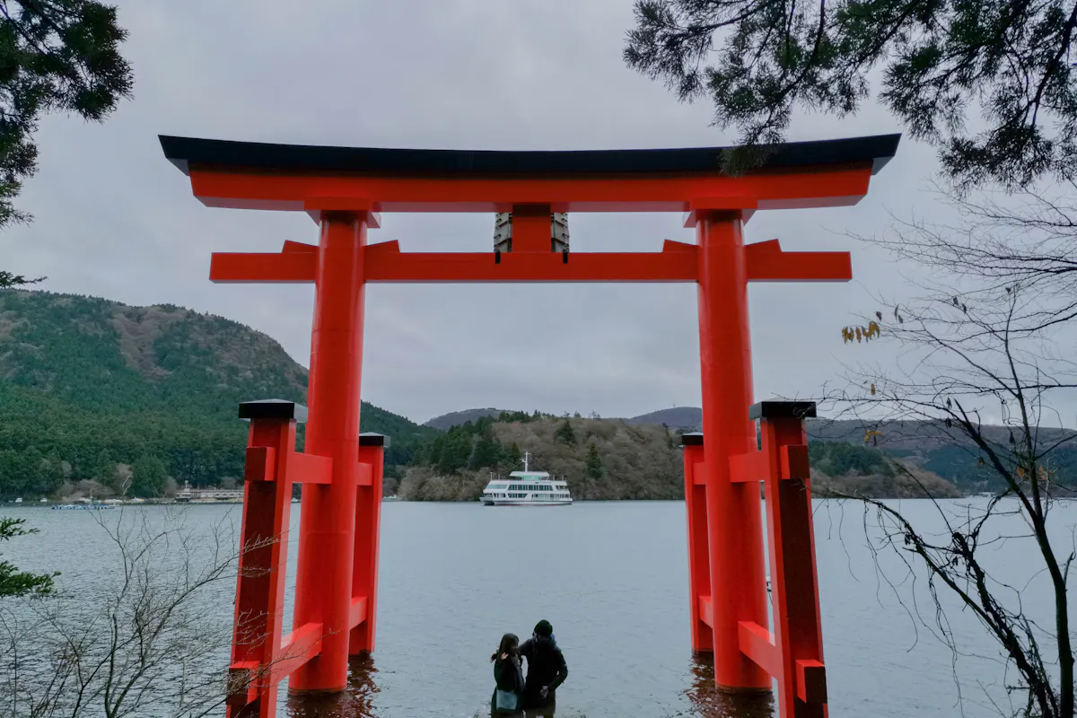 Hakone Shrine A large red torii gate stands in a serene lakeside setting. Two people sit close to the water, looking at a ferry in the distance. Surrounding the scene are lush green mountains and a cloudy sky.