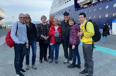 A group of eight people, dressed in casual jackets and backpacks, stand and smile together on a pier next to a large cruise ship. The weather looks cool and clear.