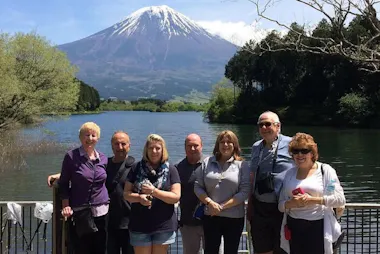 A group of seven people pose together by a lake with snow-capped Mount Fuji in the background, surrounded by trees and greenery on a clear, sunny day.