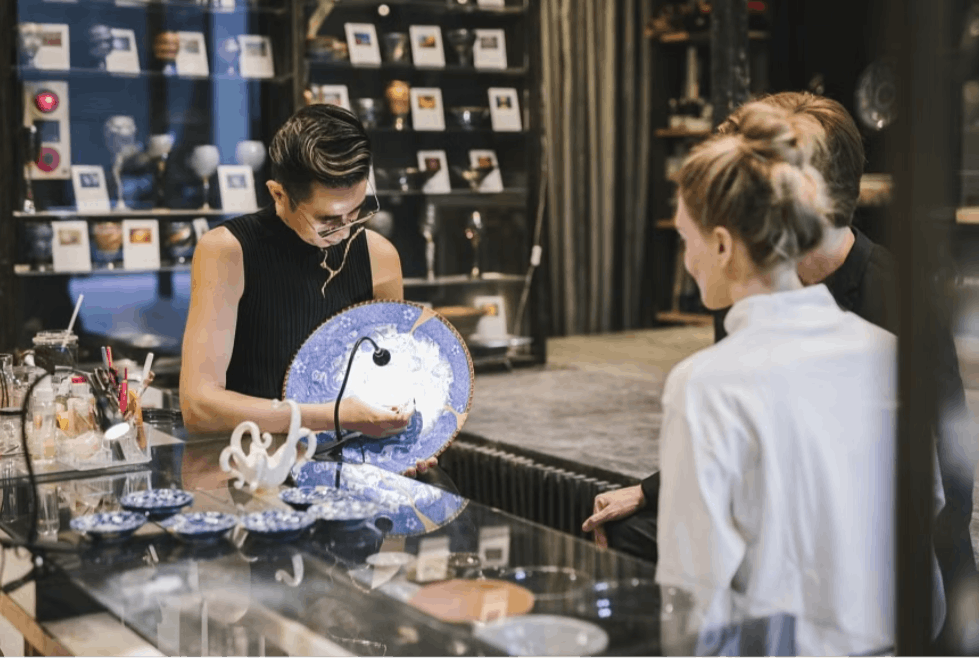 A person stands behind a glass counter, showing a large blue and white ceramic plate to two customers in a shop filled with pottery, ceramics, and art pieces.
