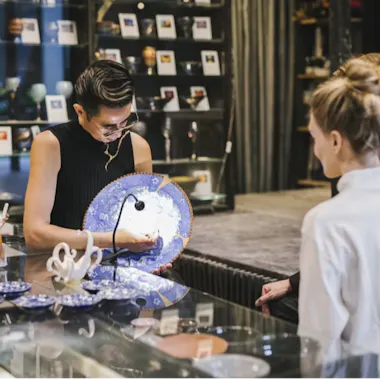 Kintsugi A person stands behind a glass counter, showing a large blue and white ceramic plate to two customers in a shop filled with pottery, ceramics, and art pieces.