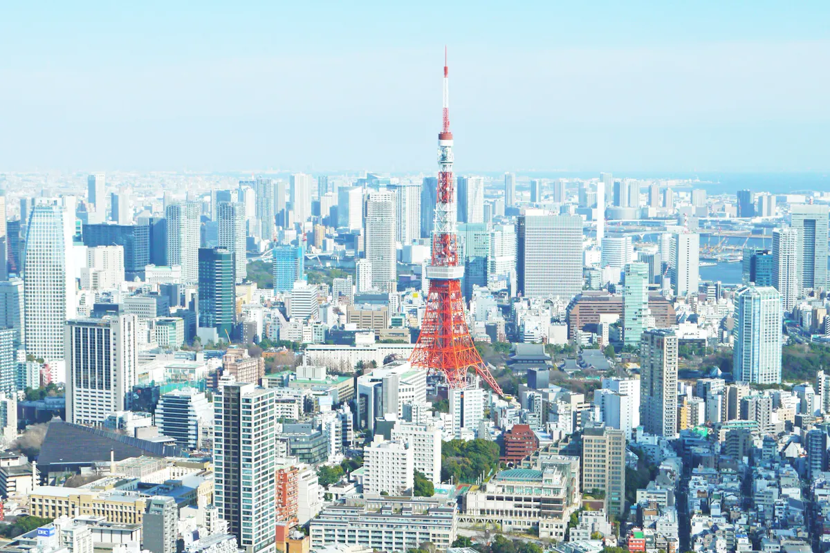 Tokyo Tower A panoramic view of Tokyo with the iconic Tokyo Tower in the center. Surrounding the tower are numerous high-rise buildings and skyscrapers under a clear blue sky, with the horizon visible in the background.