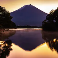 Lake Tanuki Snow-capped mountain reflected in a calm lake at sunrise, surrounded by trees with a golden sky creating a tranquil and scenic landscape.