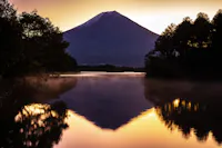 Snow-capped mountain reflected in a calm lake at sunrise, surrounded by trees with a golden sky creating a tranquil and scenic landscape.