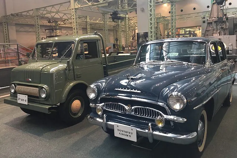 An indoor exhibit showcases two classic vehicles: a green, vintage truck on the left labeled "PR10-N" and a dark blue, vintage car on the right labeled "Toyopet Crown." The vehicles are parked side by side amidst an industrial backdrop with metal beams.