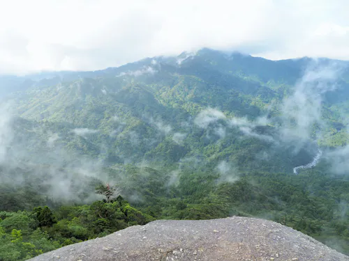 View from a rocky ledge overlooking lush green mountains partially covered by mist and low clouds, with a river visible in the distance under a bright, cloudy sky.