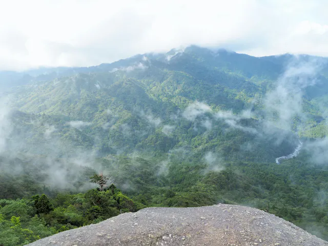 View from a rocky ledge overlooking lush green mountains partially covered by mist and low clouds, with a river visible in the distance under a bright, cloudy sky.
