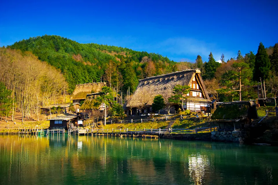Hida Folk Village 穏やかな風景、伝統的な茅葺き屋根の家々が木々の間に立ち、反射する澄んだ湖のそばに位置しています。前景には穏やかな水面が広がり、背景には明るく澄んだ青空の下に豊かな緑の森が広がっています。