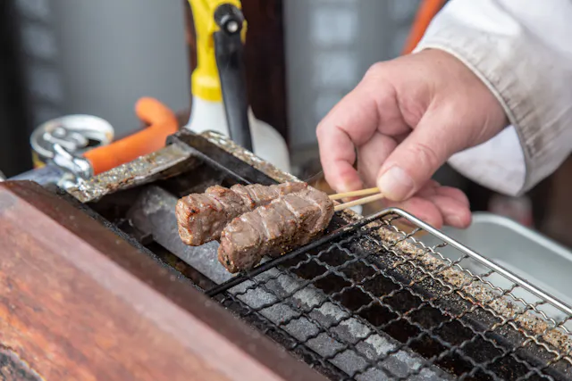 A person grilling two skewers of meat over a metal grate, using their hand to turn the skewers. A spray bottle and other utensils are visible in the background.