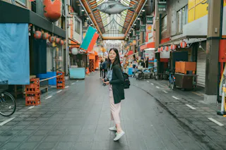 A young woman with a backpack stands smiling in the middle of a covered shopping street adorned with lanterns and a large fish decoration overhead. She holds a camera and looks back at the camera. The street appears calm and mostly empty.