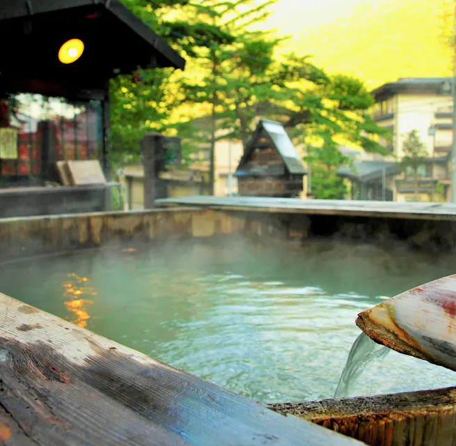 A serene outdoor hot spring bath with warm, steaming water flowing from a wooden spout. Surrounded by greenery and traditional wooden buildings, it creates a peaceful and relaxing atmosphere against the backdrop of a mellow sunset.