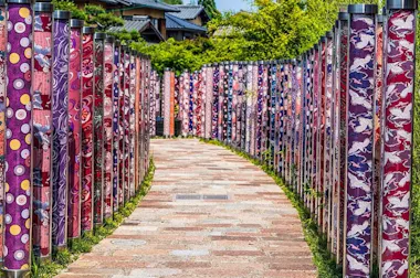 A winding stone path lined with tall cylindrical pillars covered in colorful, patterned fabric, featuring reds, purples, and designs of birds and geometric shapes. Lush greenery and buildings are visible in the background.