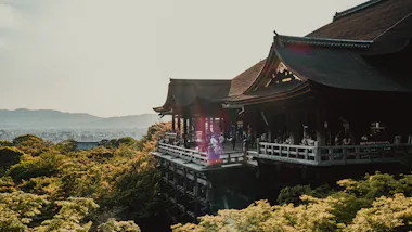 A large traditional Japanese wooden temple sits elevated above lush green trees with people on its balcony, overlooking a hazy city and distant mountains under a bright sky.