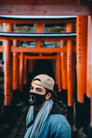 A person wearing a beige cap, a scarf, and a black face mask with a peace sign stands in front of a series of red torii gates at a shrine, looking upward.