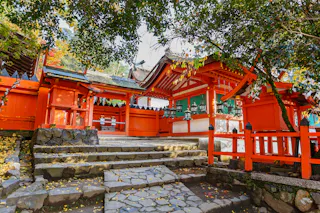 A traditional Japanese Shinto shrine with bright red wooden structures, stone steps, and surrounding trees, illuminated by natural daylight.