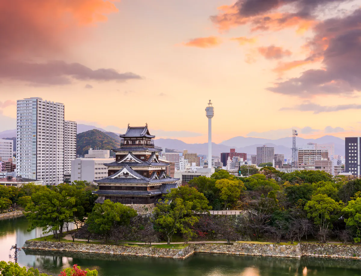 Hiroshima Castle and Cityscape A traditional Japanese castle surrounded by trees and a moat stands against a backdrop of modern city buildings and a sunset sky with orange clouds in Hiroshima, Japan.