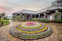 A circular flower bed with colorful blooms is centered on a stone walkway in front of a historic house with a tiled roof, arched veranda, and pastel shutters under a cloudy sky.