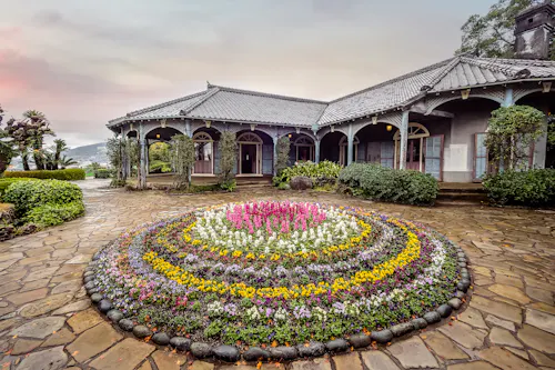 A circular flower bed with colorful blooms is centered on a stone walkway in front of a historic house with a tiled roof, arched veranda, and pastel shutters under a cloudy sky.