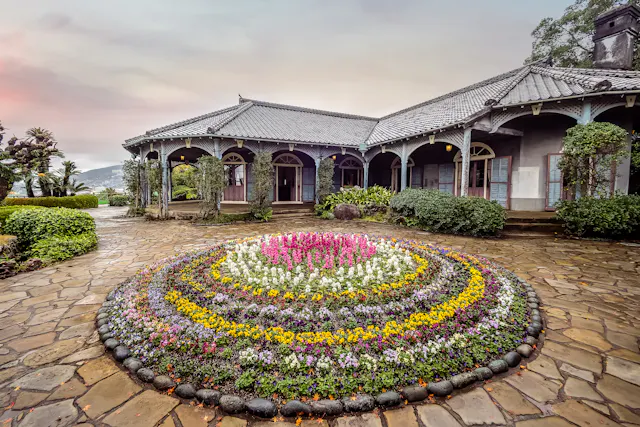 A circular flower bed with colorful blooms is centered on a stone walkway in front of a historic house with a tiled roof, arched veranda, and pastel shutters under a cloudy sky.