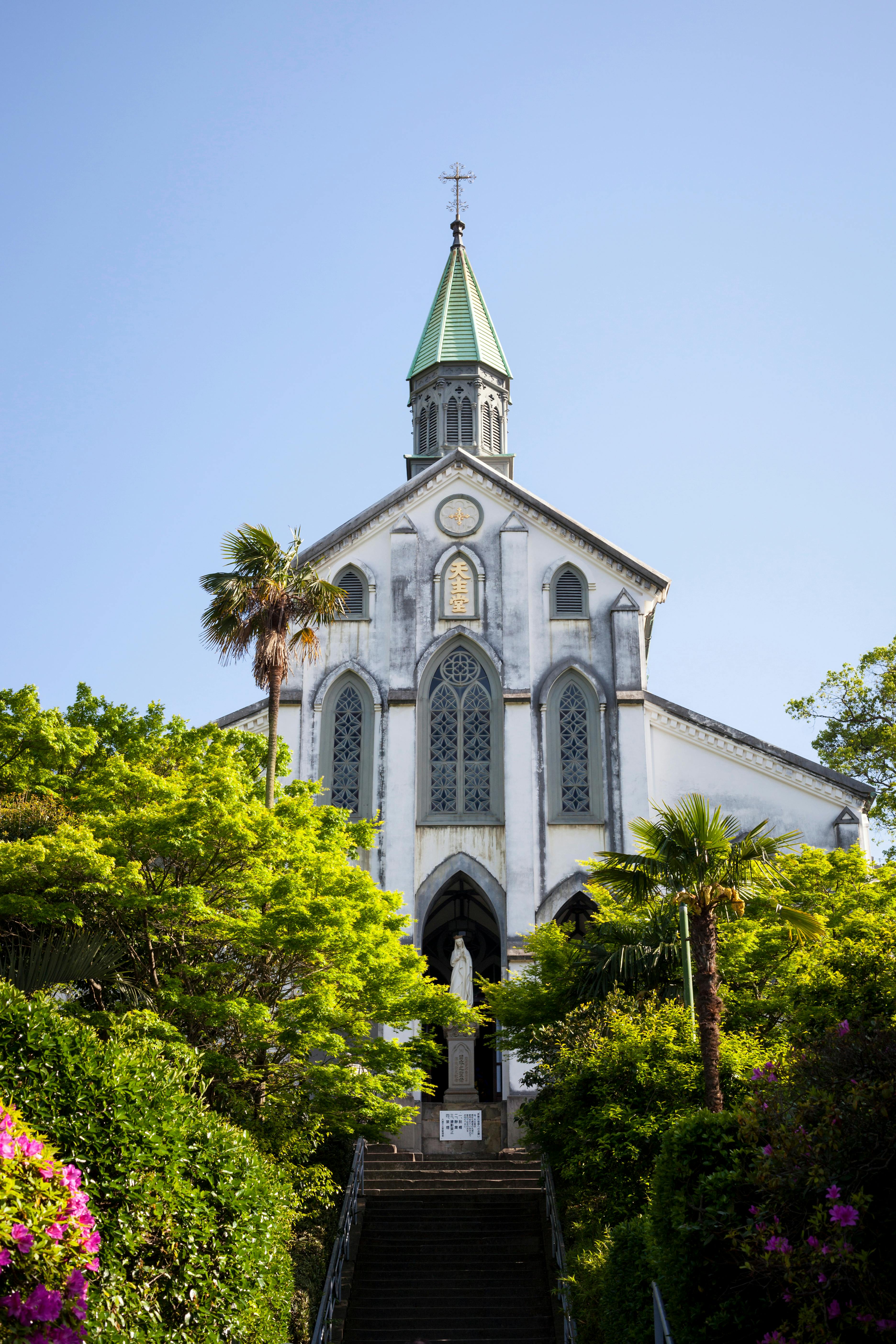 A white church with tall arched windows and a green spire stands atop a staircase, surrounded by lush green trees and colorful flowers under a clear blue sky.