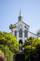 A white church with tall arched windows and a green spire stands atop a staircase, surrounded by lush green trees and colorful flowers under a clear blue sky.