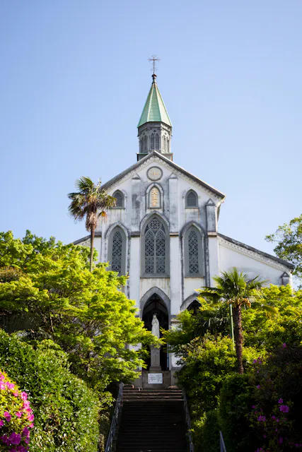 A white church with tall arched windows and a green spire stands atop a staircase, surrounded by lush green trees and colorful flowers under a clear blue sky.