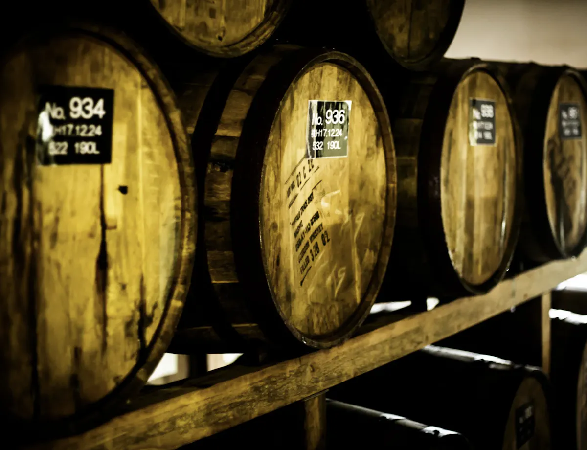 Brewery A row of wooden barrels, each with identification numbers and capacity details, are stored on wooden shelves in a dimly lit room. The barrels appear aged, suggesting they hold wine, whiskey, or another type of spirits.