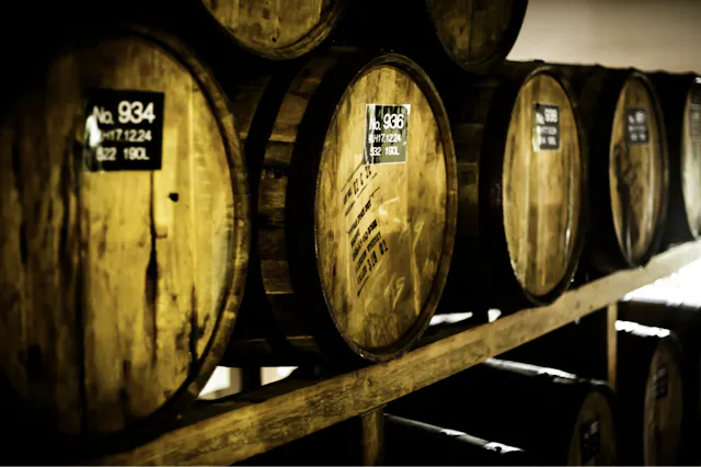 A row of wooden barrels, each with identification numbers and capacity details, are stored on wooden shelves in a dimly lit room. The barrels appear aged, suggesting they hold wine, whiskey, or another type of spirits.