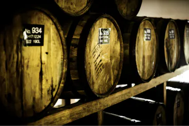 A row of wooden barrels, each with identification numbers and capacity details, are stored on wooden shelves in a dimly lit room. The barrels appear aged, suggesting they hold wine, whiskey, or another type of spirits.