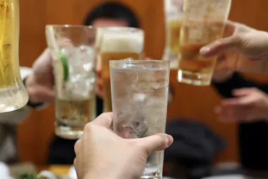 A group of people raise their glasses in a toast, clinking together various beverages, including clear and amber drinks, against a blurred indoor background.