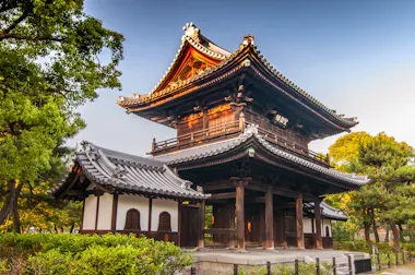 A traditional Japanese wooden temple gate with curved tiled roofs stands surrounded by lush green trees, lit by warm sunlight under a clear blue sky.