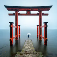 Hakone Shrine A person holding a white umbrella stands on a stone path leading to a large red torii gate over calm water, surrounded by mist and soft light.