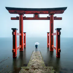 Hakone Shrine A person holding a white umbrella stands on a stone path leading to a large red torii gate over calm water, surrounded by mist and soft light.