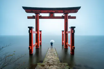 Hakone Shrine A person holding a white umbrella stands on a stone path leading to a large red torii gate over calm water, surrounded by mist and soft light.