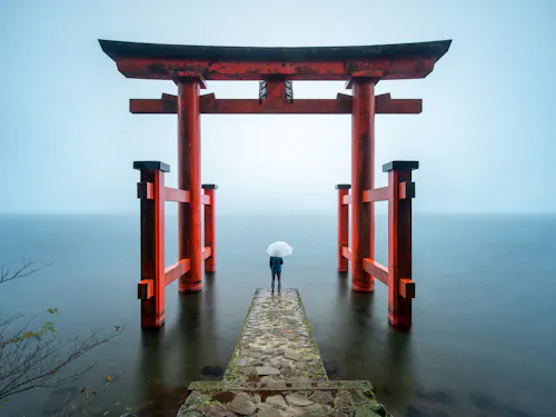 Hakone Shrine A person holding a white umbrella stands on a stone path leading to a large red torii gate over calm water, surrounded by mist and soft light.