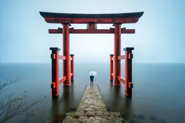 A person holding a white umbrella stands on a stone path leading to a large red torii gate over calm water, surrounded by mist and soft light.