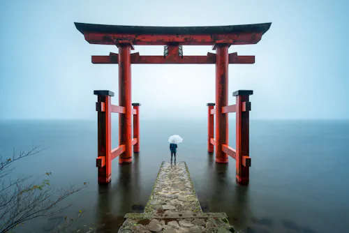 A person holding a white umbrella stands on a stone path leading to a large red torii gate over calm water, surrounded by mist and soft light.