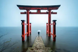 A person holding a white umbrella stands on a stone path leading to a large red torii gate over calm water, surrounded by mist and soft light.