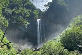 A tall waterfall cascades down a rocky cliff surrounded by lush green trees and vegetation, with mist rising at the base and sunlight streaming through the scene.