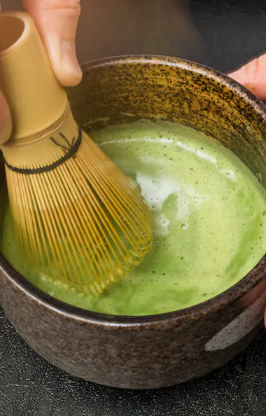 Tea Ceremony A person uses a bamboo whisk to mix frothy green matcha tea in a dark, ceramic bowl on a black surface.