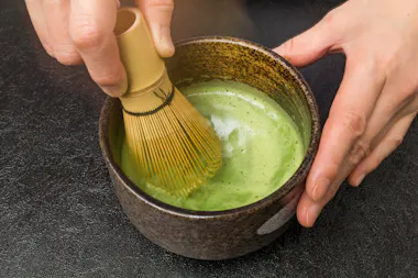 A person uses a bamboo whisk to mix frothy green matcha tea in a dark, ceramic bowl on a black surface.