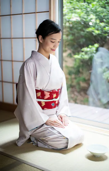 Tea Ceremony A woman wearing a light-colored kimono with a red obi sits on a tatami mat by a window, hands resting on her knees, with a tea bowl in front of her and greenery visible outside.