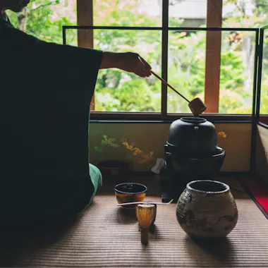 Tea Ceremony Camellia FLOWER A person in traditional Japanese clothing prepares tea on a tatami mat, using a ladle, teapot, and tea bowls, with a garden visible through the window in the background.