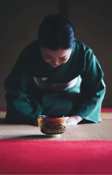 Tea Ceremony Camellia FLOWER A woman in a green kimono kneels on a tatami mat, bowing respectfully with a traditional tea bowl in front of her, inside a softly-lit Japanese room.