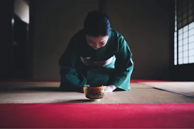 A woman in a green kimono kneels on a tatami mat, bowing respectfully with a traditional tea bowl in front of her, inside a softly-lit Japanese room.
