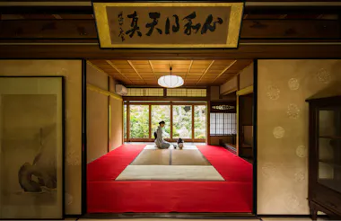A person in traditional Japanese clothing sits on a red mat in a tatami room, facing a tea set. The room has sliding doors and opens to a garden view, with soft natural light illuminating the scene.