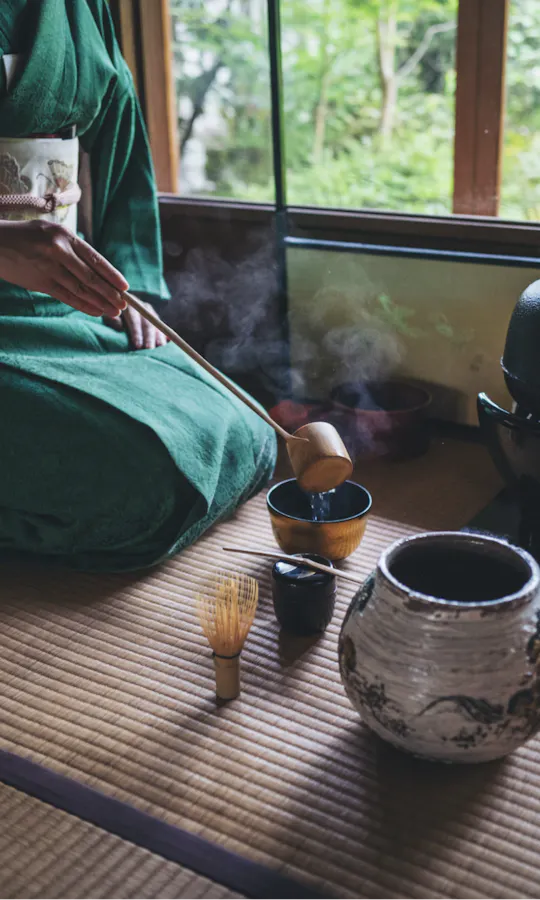 Tea Ceremony Camellia FLOWER A person in a green kimono prepares tea on a tatami mat, using traditional Japanese tea ceremony utensils, with steam rising and a garden visible through the window.