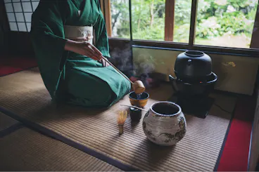 A person in a green kimono prepares tea on a tatami mat, using traditional Japanese tea ceremony utensils, with steam rising and a garden visible through the window.