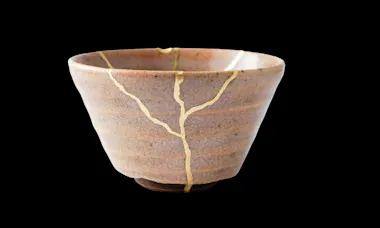 A ceramic bowl repaired with visible gold lines along its cracks, highlighting the Japanese art of kintsugi, set against a solid black background.